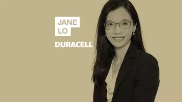 Person in a formal outfit with glasses, smiling against a solid background.