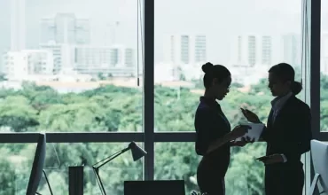Two colleagues discussing work by an office window with a city view.