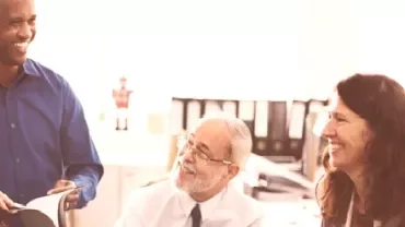 Three people smiling and discussing in an office setting.